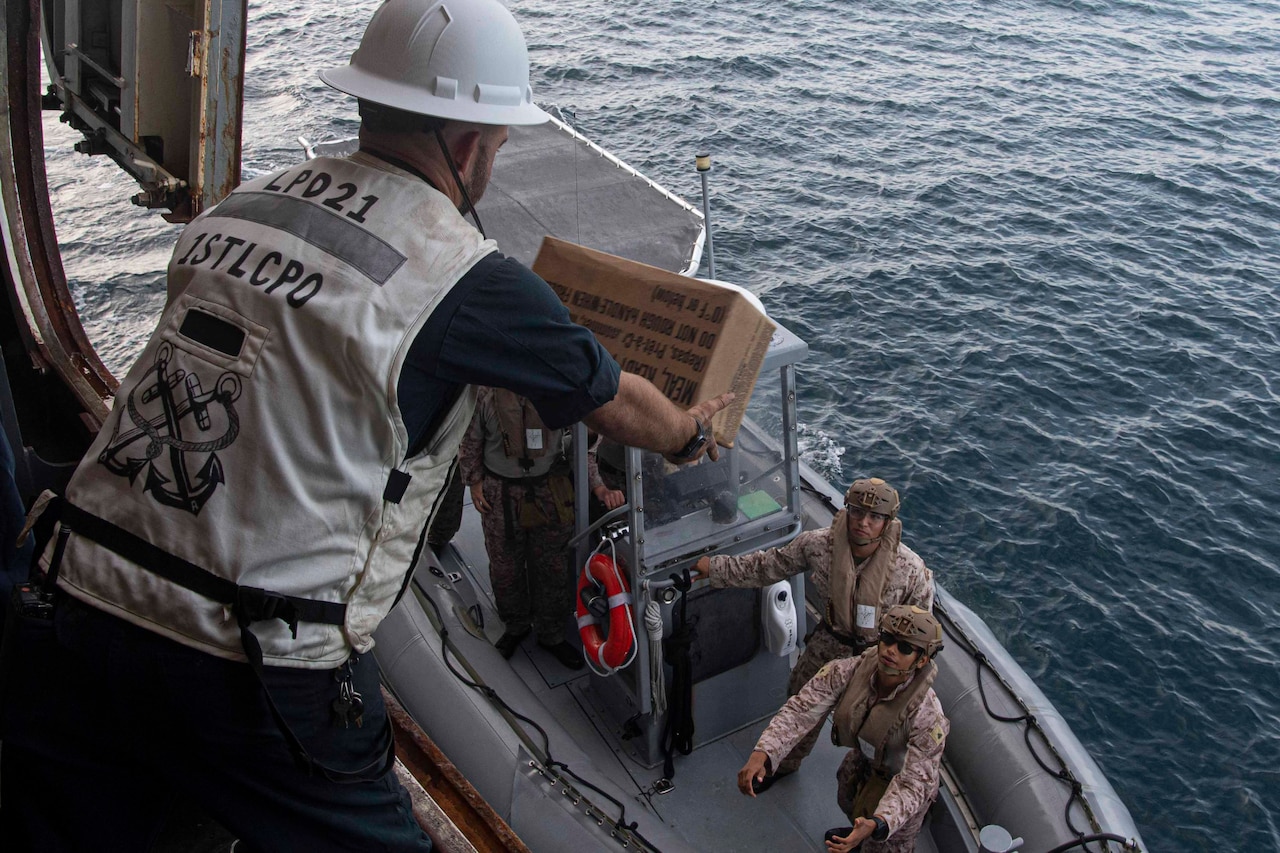 A sailor aboard a ship prepares to toss a box to Marines in a small inflatable boat.