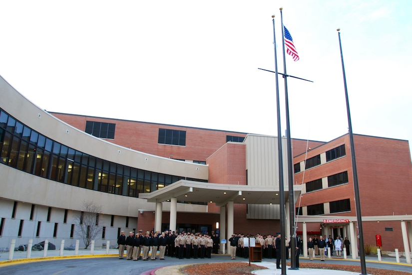 A group of sailors in uniform stand in formation as a sailor raises the American flag outside of a building
