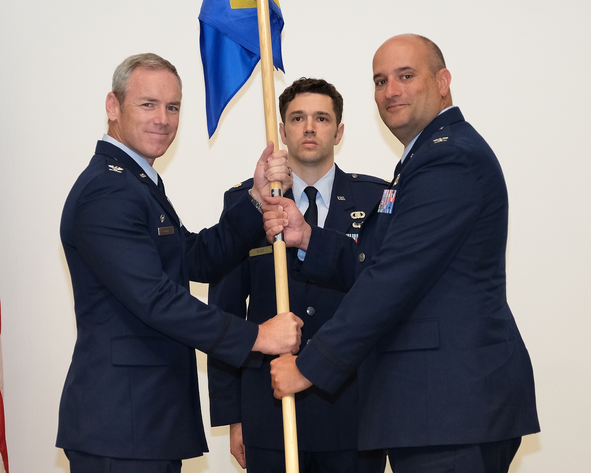 photo of three uniformed US Air Force Airmen standing on a stage, two Airmen in the forefront are both holding a unit flag while third Airman stands in background at attention