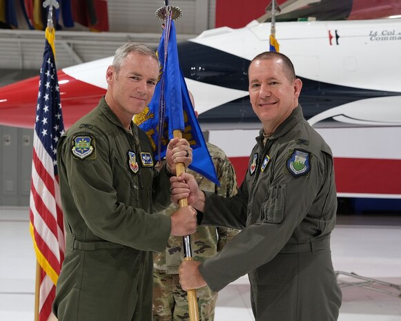 photo of three uniformed US Air Force Airmen standing on a stage, two Airmen in the forefront are both holding a unit flag while third Airman stands in background at attention