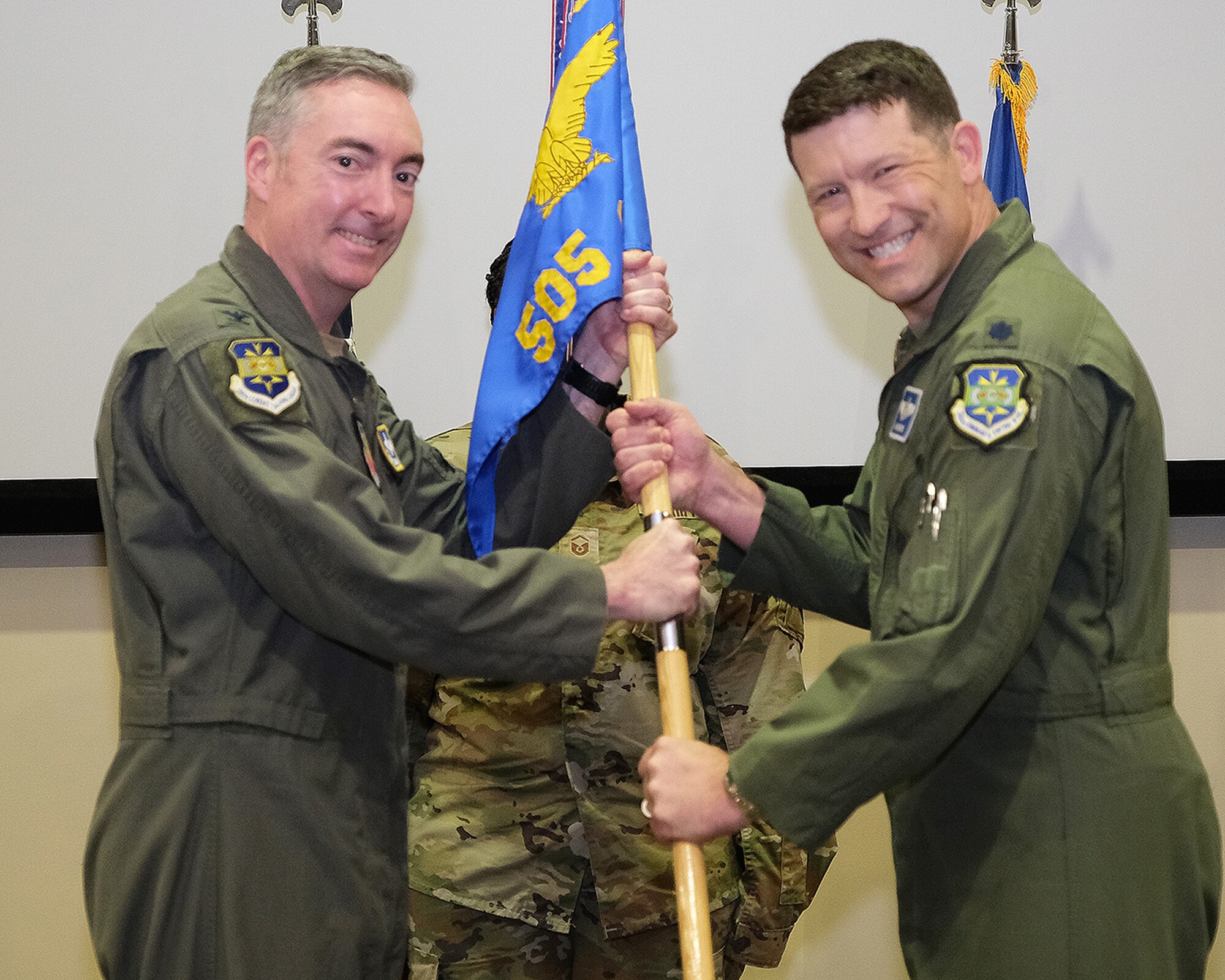 photo of three uniformed US Air Force Airmen standing on a stage, two Airmen in the forefront are both holding a unit flag while third Airman stands in background at attention