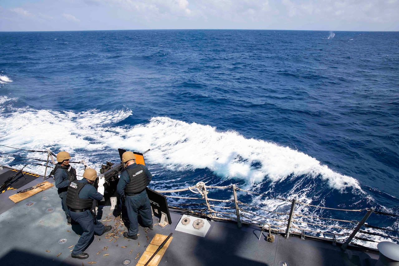 Sailors shoot a gun from the deck of a ship as the ship travels through a body of water.