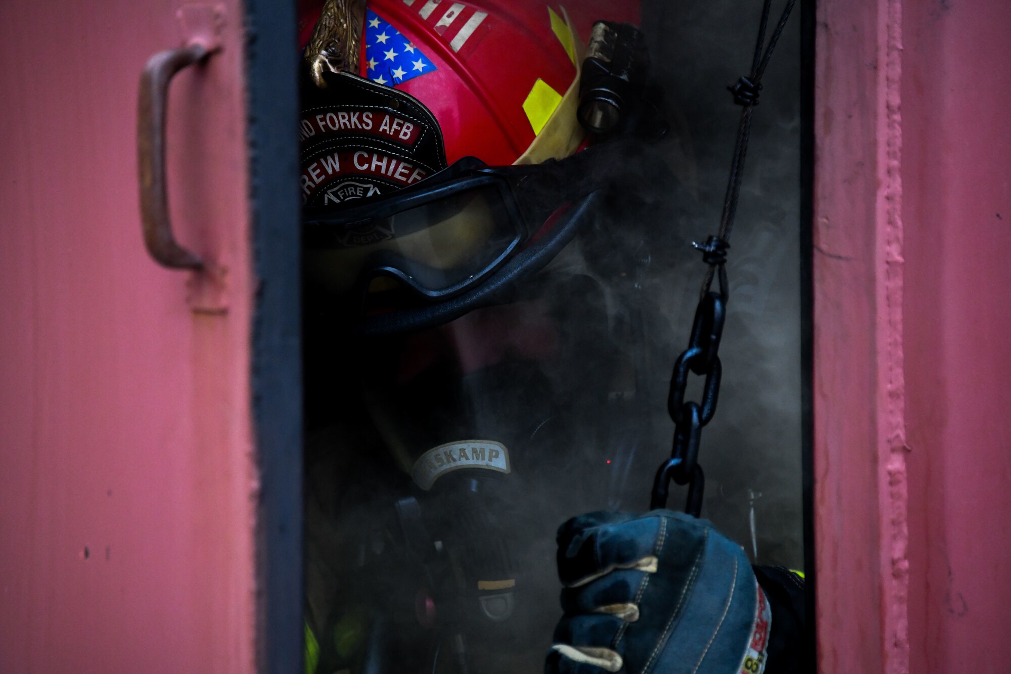 A man in a firefighter uniform sticks his head out of a door with smoke all around him.