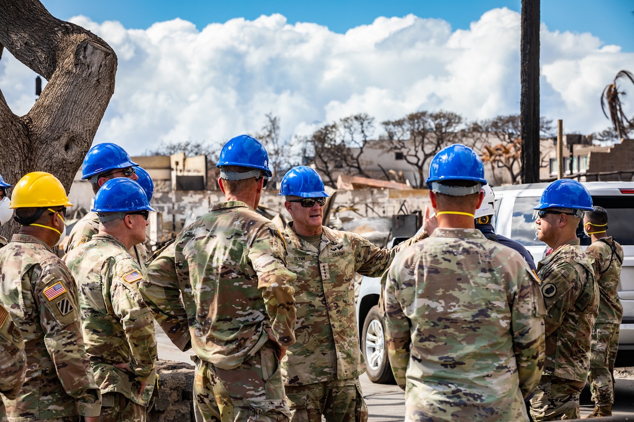 Uniformed soldiers talk while wildfire-damaged structured can be seen in the background.