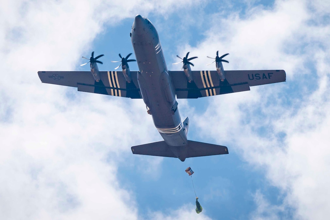 A look underneath an airborne aircraft as a package attached to a parachute descends in the sky below it.