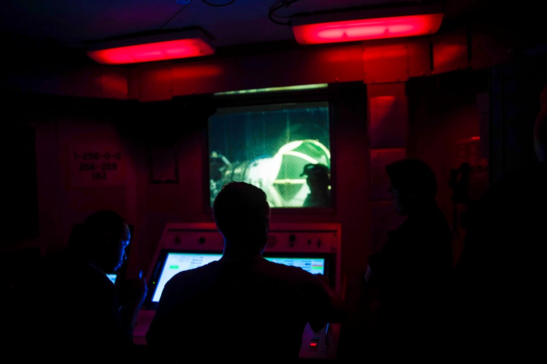 Sailors sit in front of computer monitors in a dimly lit room.