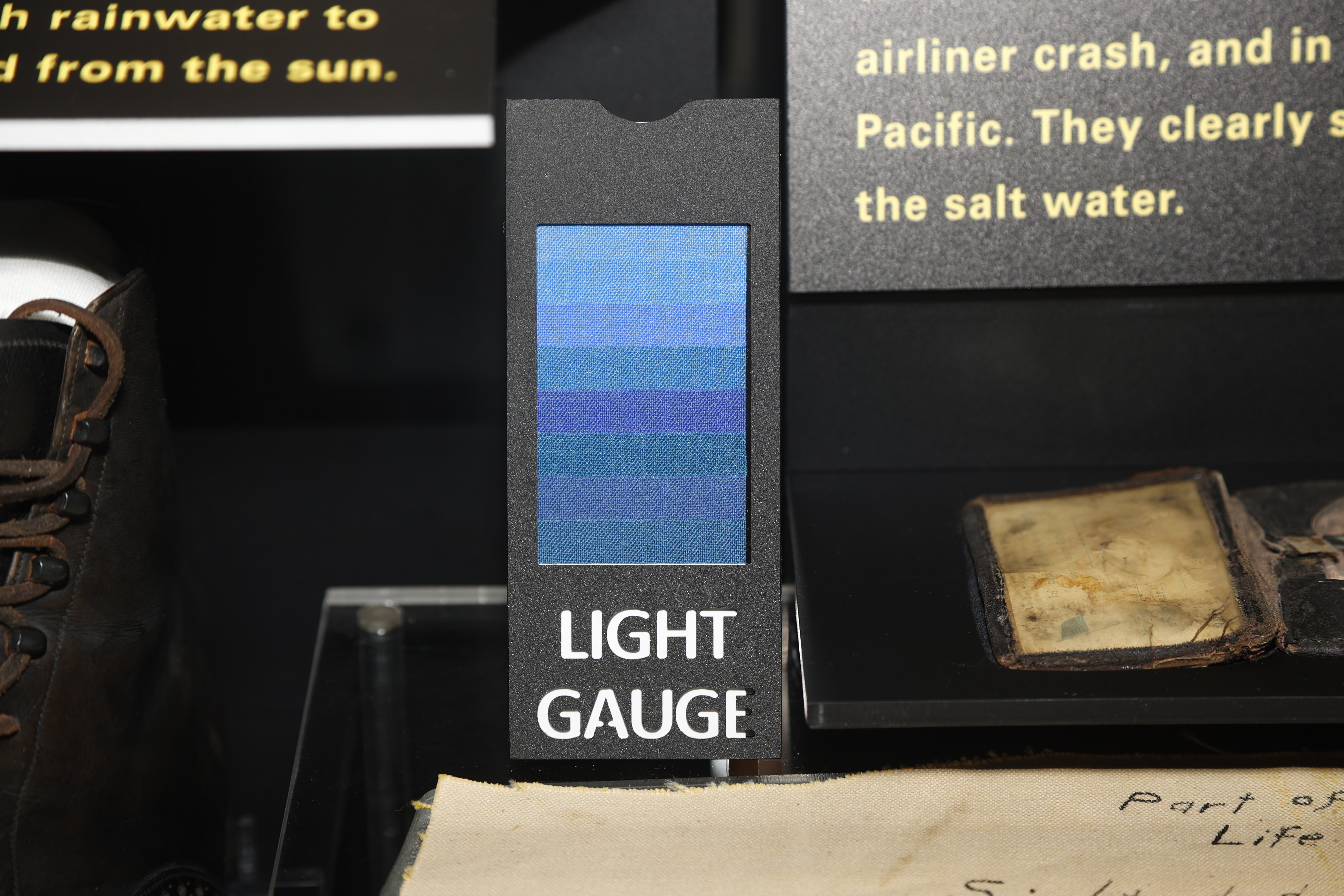 어두운 박물관 진열장 안에서 여덟 단계의 파란 천 조각이 붙은 검은색 LIGHT GAUGE 표지가 낡은 종이 유물 옆에 세워진 모습