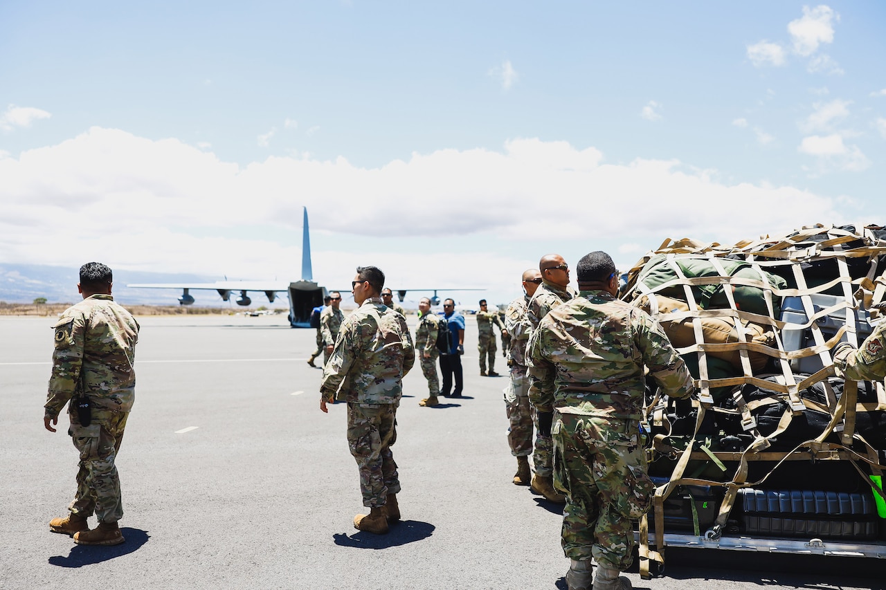 Service members load cargo on a tarmac with a military aircraft in the distance.