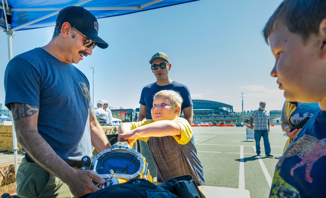 Navy Diver Petty Officer 1st Class Matthew Villafuerte, Keyport Dive Locker supervisor, and Navy Diver Petty Officer 1st Class Christian Stewart, Everett Dive Locker supervisor, watch as a curious Seafair visitor tries to lift a 33-pound diving helmet Aug. 3, 2023, on Pier 46 in Seattle. (U.S Navy Photo by Wendy Hallmark)