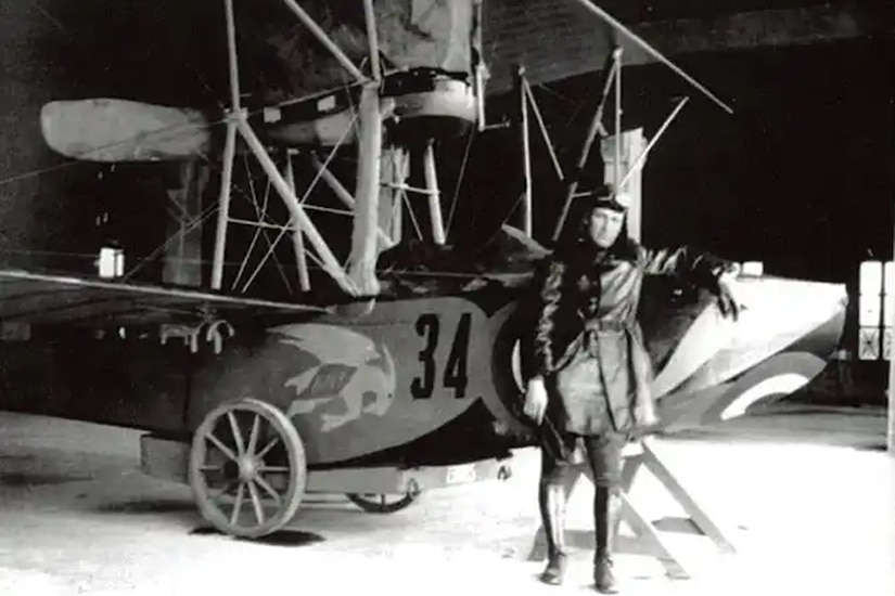 A person dressed in pilot gear stands beside a propeller airplane in a black and white photo.