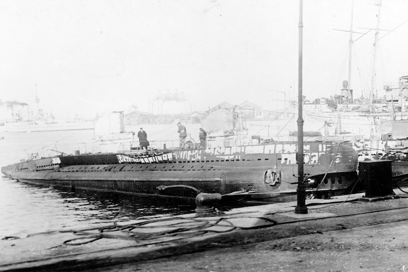 Three people stand on top of a submarine in a harbor in a black and white photo.