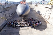 USS Tucson (SSN 770) Los Angeles-class fast-attack submarine sits in dry dock with the USS Tucson team of people standing in front of her holding a banner.