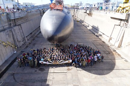 USS Tucson (SSN 770) Los Angeles-class fast-attack submarine sits in dry dock with the USS Tucson team of people standing in front of her holding a banner.