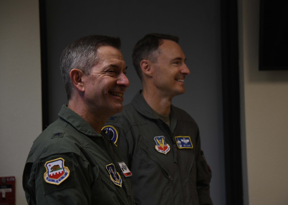 Two men in military uniforms smile and face off to right to something out of the frame.