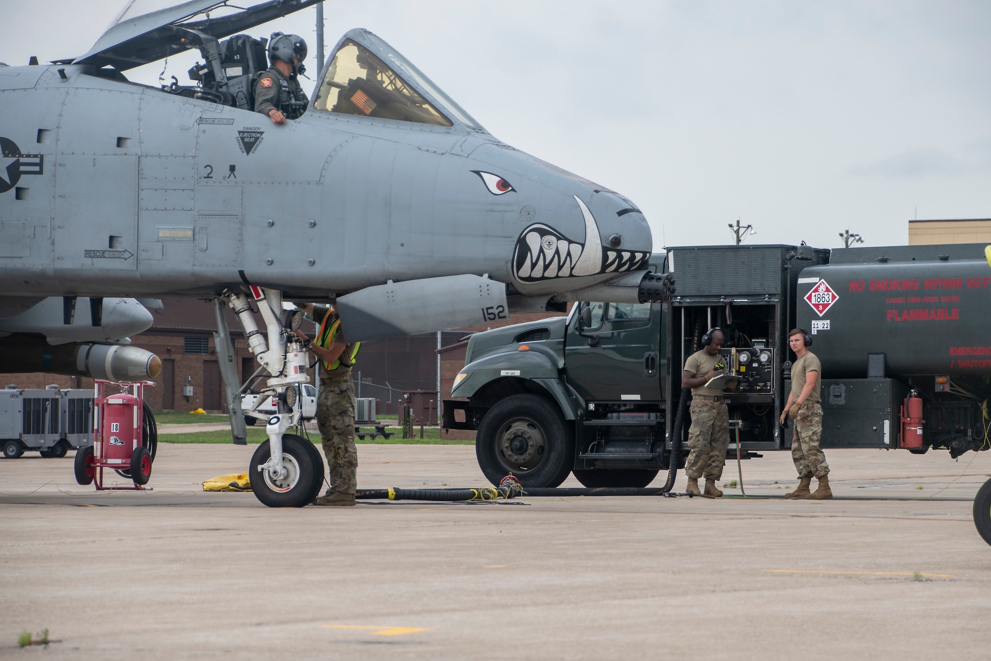 Airmen in camo uniforms prepare to pump gas from a green truck into a gray fighter aircraft with teeth painted on nose.
