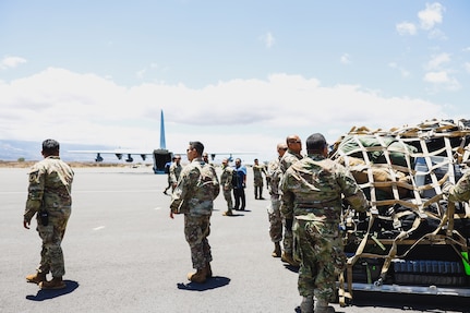Service members load cargo on a tarmac with a military aircraft in the distance.