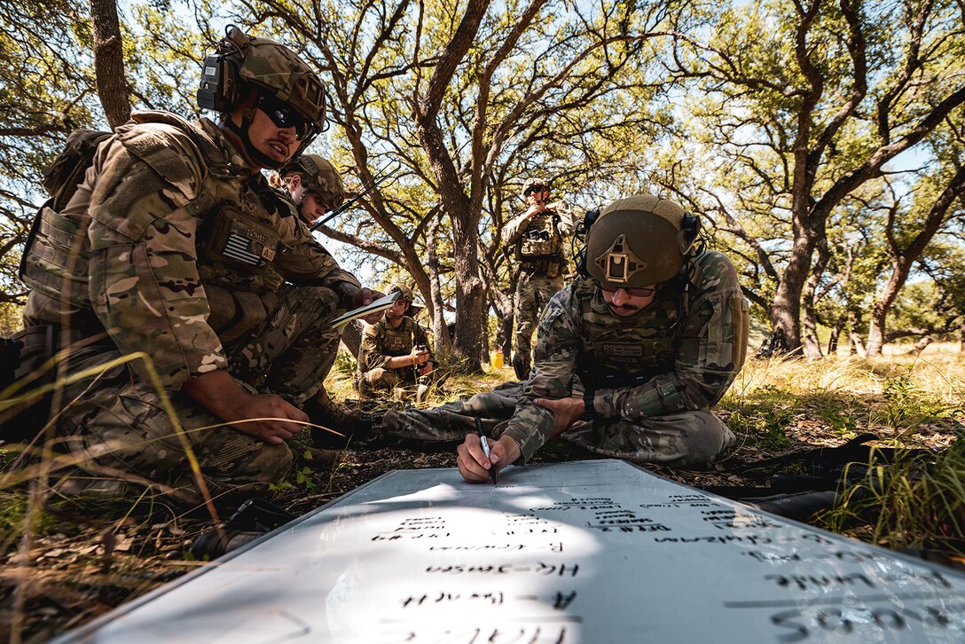 A uniformed service member writes on a dry erase board placed on the grass as others look on.