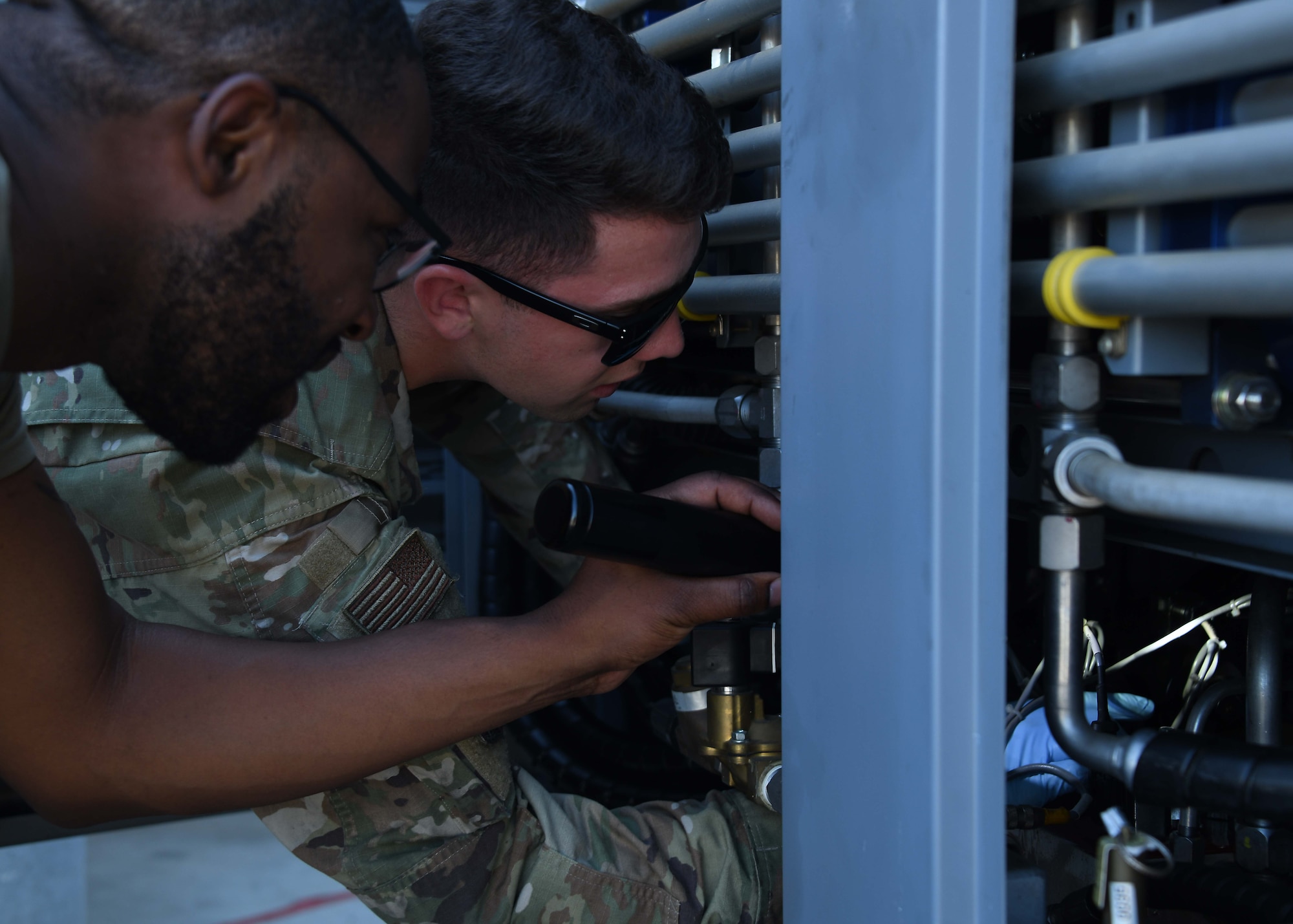 From the left, two US service members look into a gray metallic machine with a flashlight.