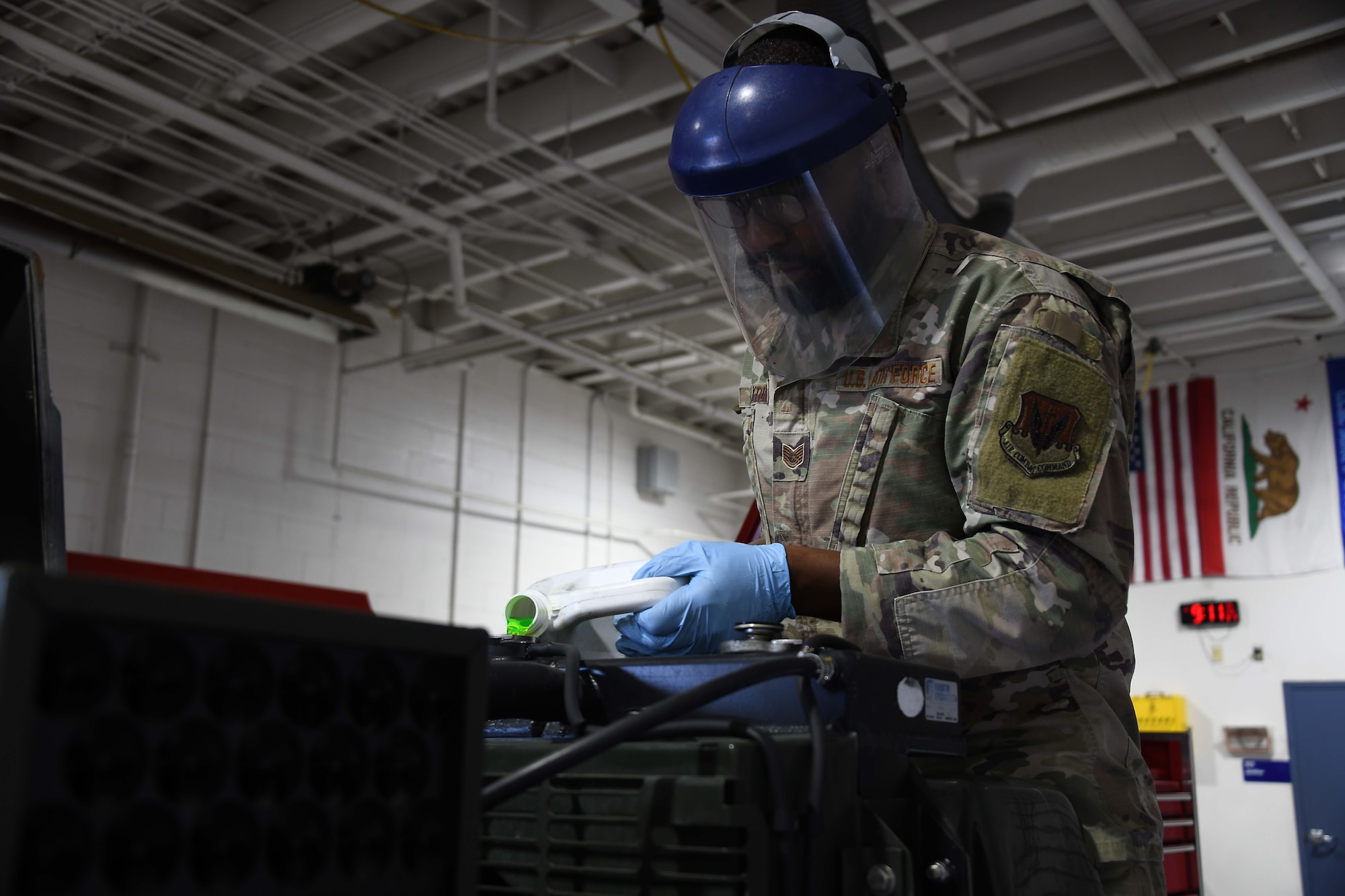 A US service member wearing green camo, a face shield and blue gloves pours green fluid into a metallic machine.