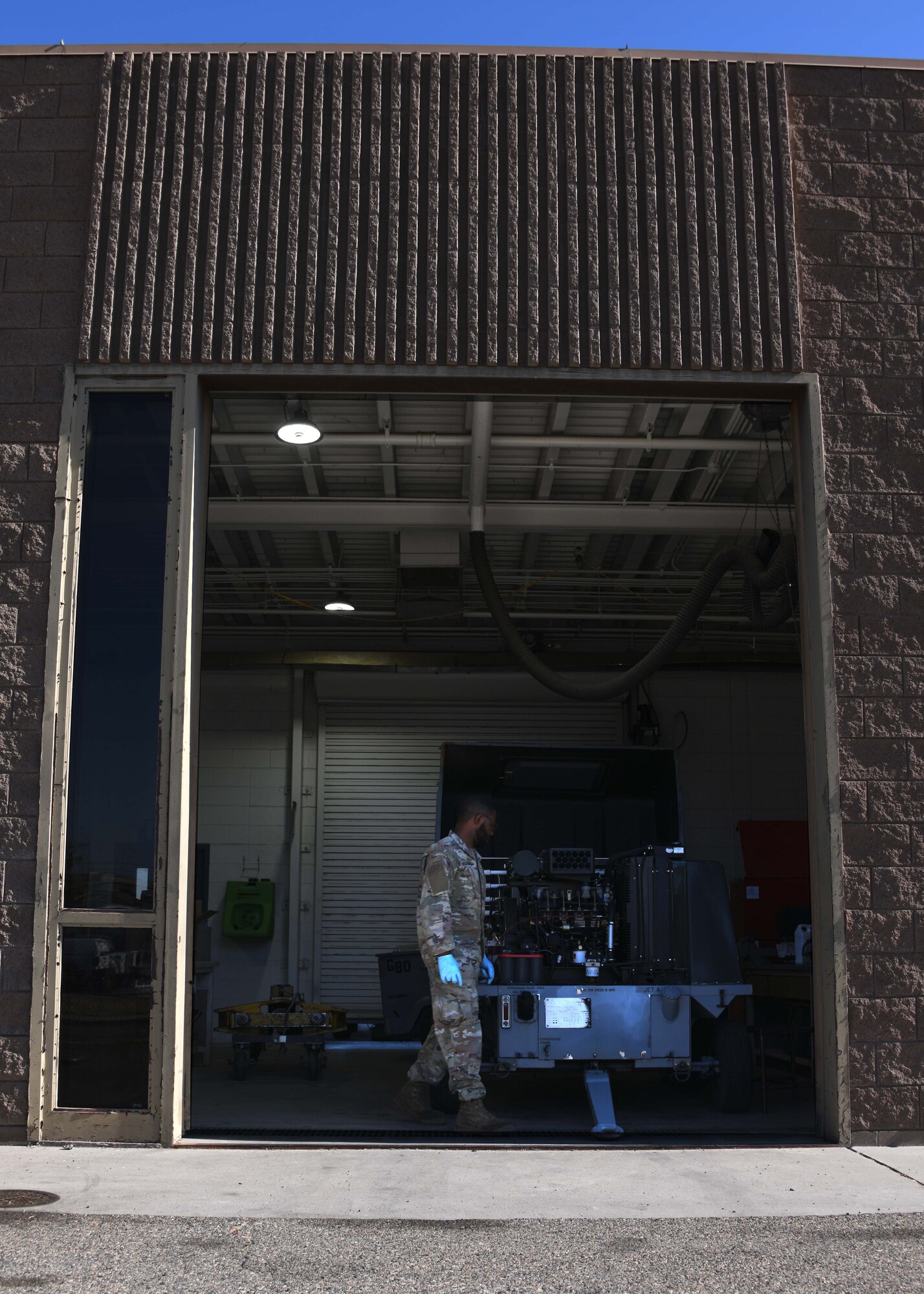 Seen from the outside of a building, A US service member wearing green camo walks around a metal machine