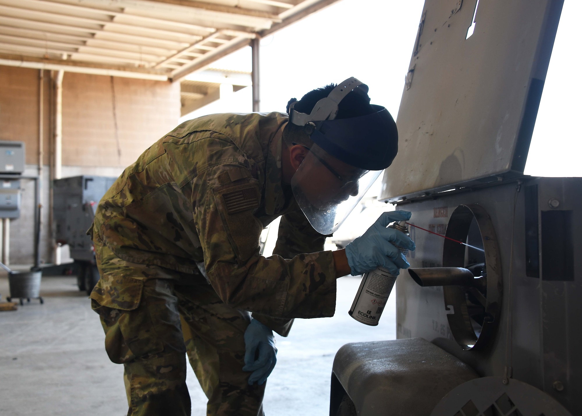 A US service member wearing green camo uses a spray can to clean the inside of a metallic machine.