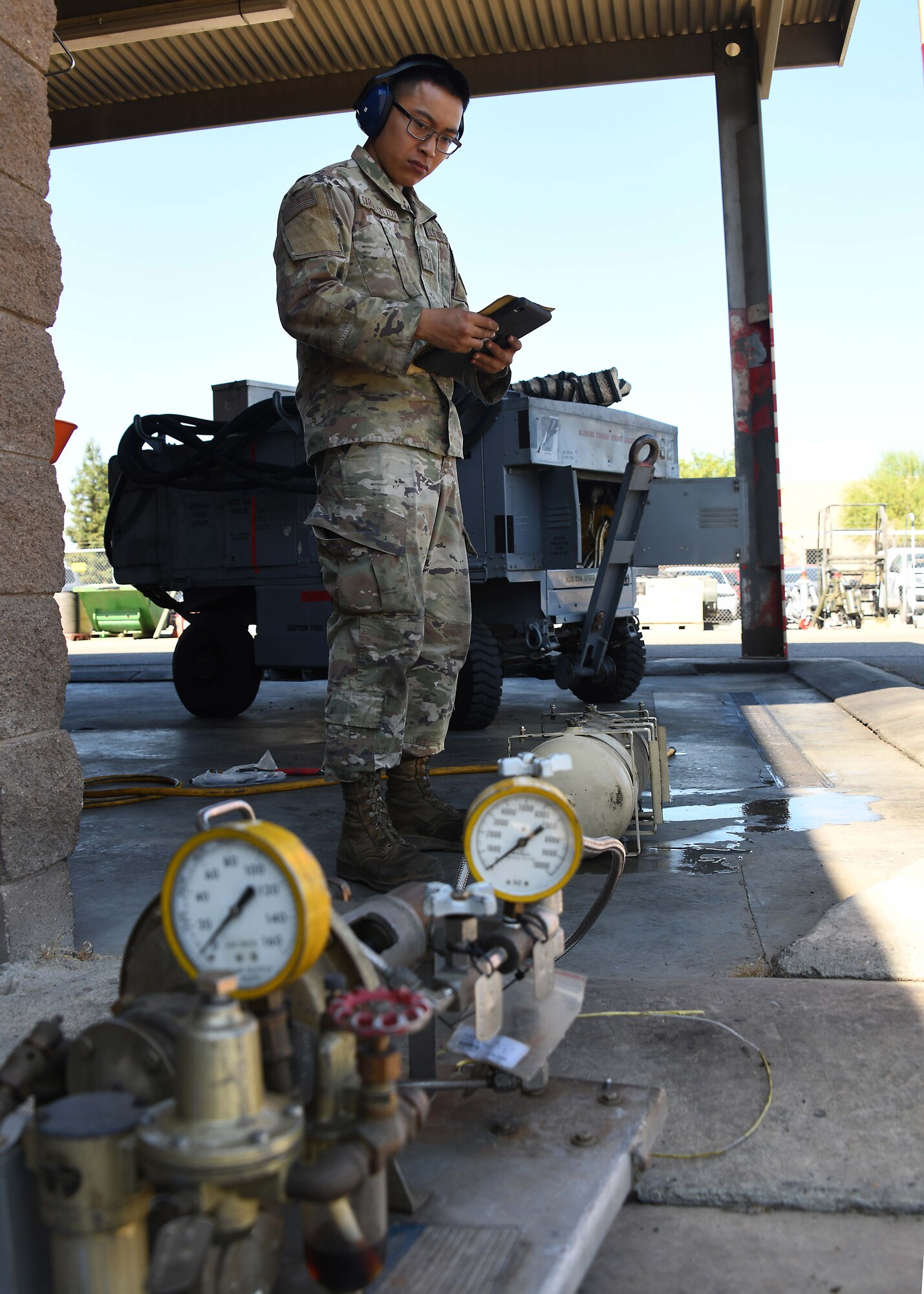 A  US service member wearing green camo reviews instructions behind two pressure gauges in the foreground.