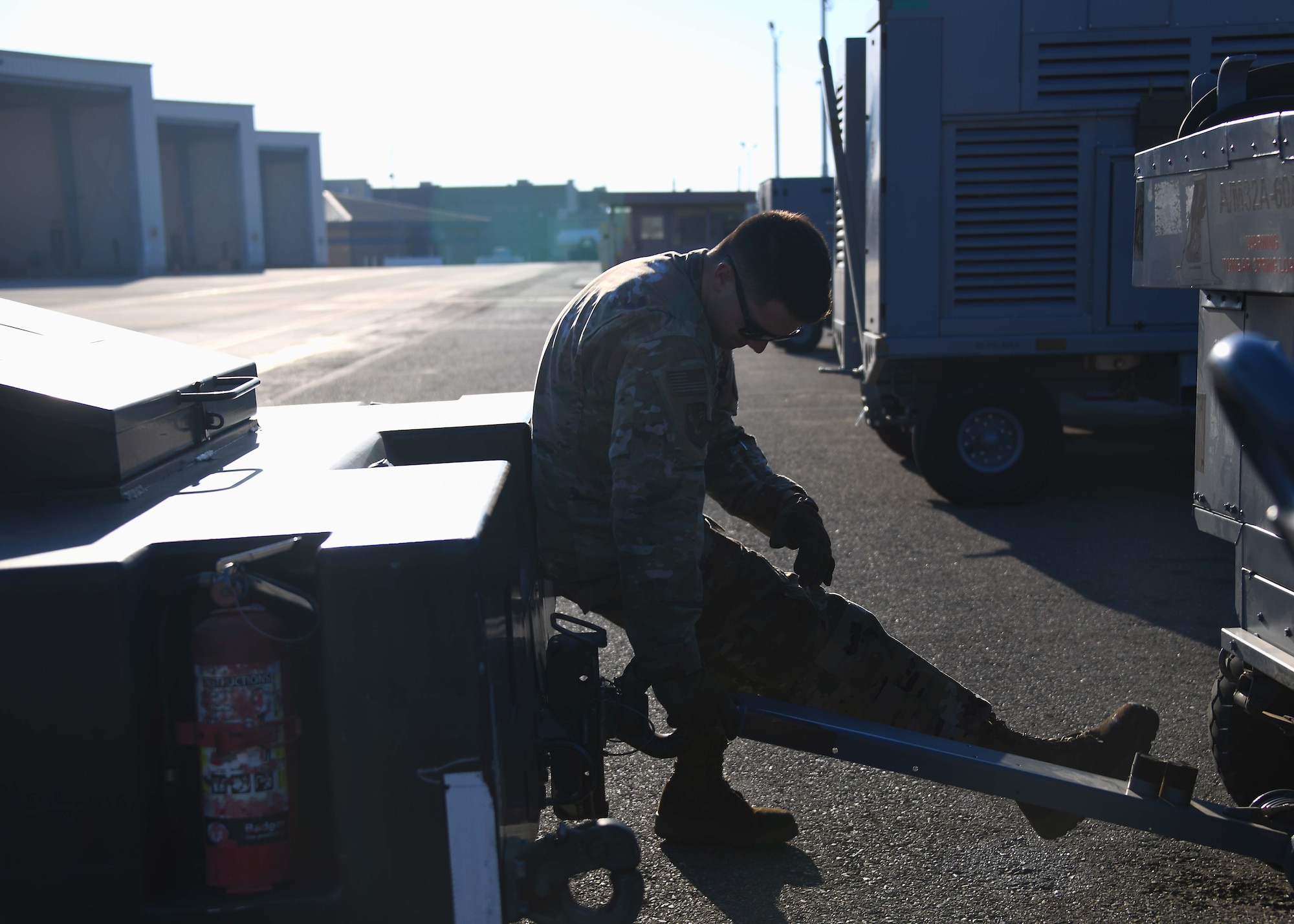 A service member wearing green camo sits on a truck while trying to attach a power generator