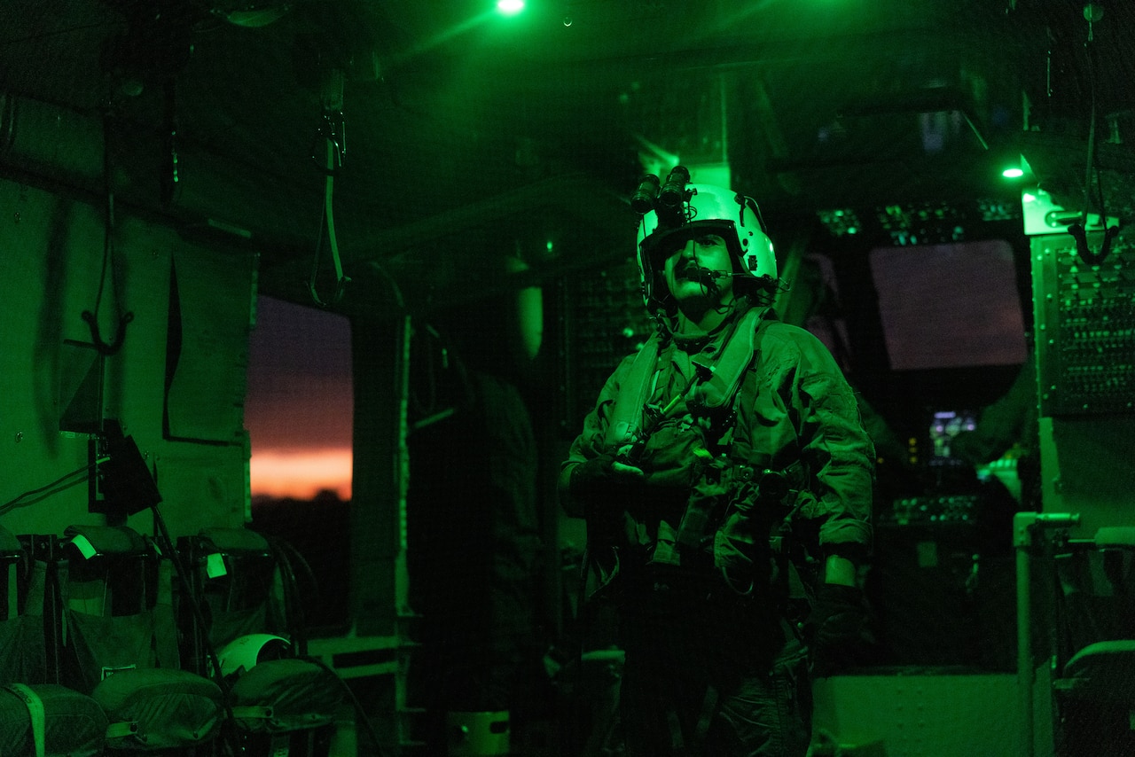 A Marine illuminated by green light stands inside of a helicopter.