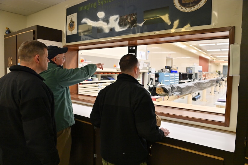 Three men observe a workroom through a window.