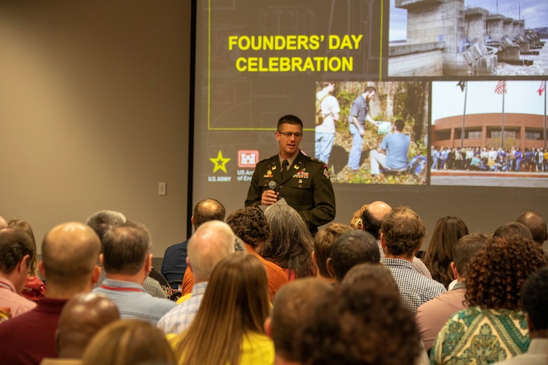 A white man in an Army colonel's uniform stands in front of a project screen that reads "Founders' Day Celebration" in bold yellow letters. In front of him are the backs of several seated peoples' heads.