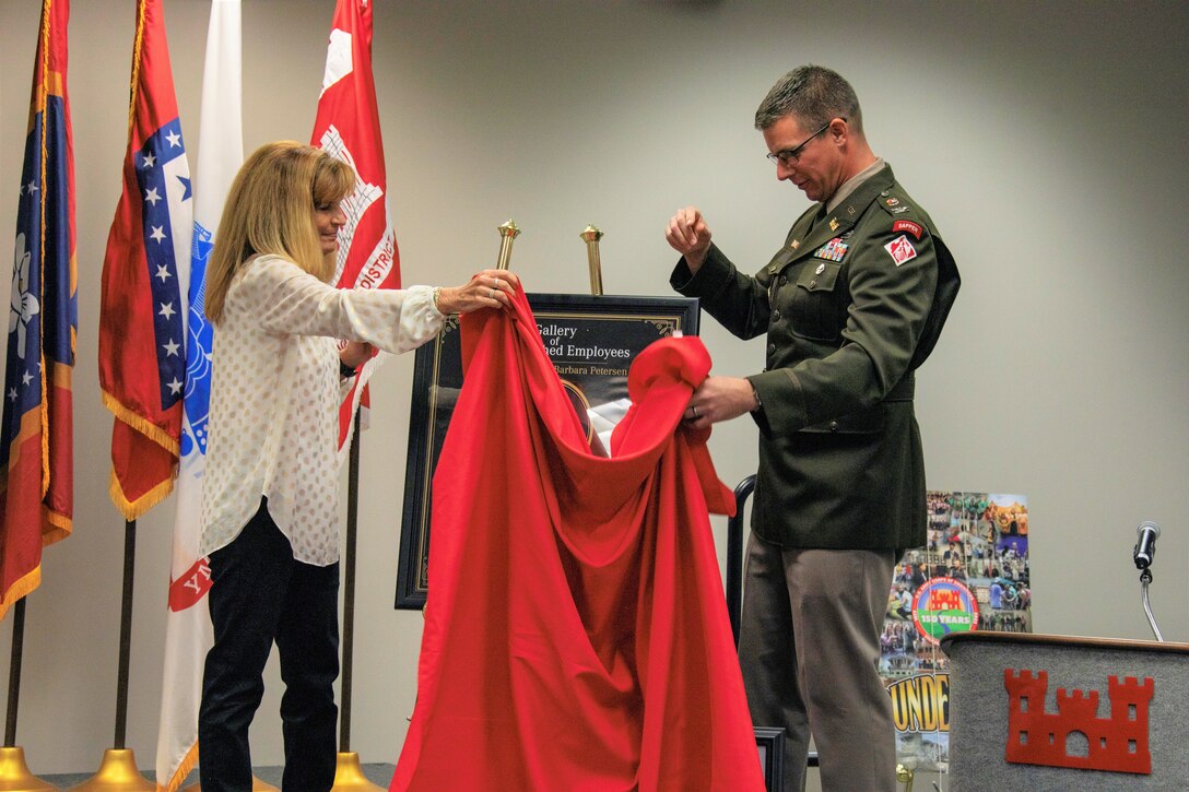 A blond-haired white woman in a white blouse and black slacks holds a red cloth between herself and a white man in an Army colonel's uniform. Behind her are the Mississippi and Arkansas state flags, the Department of the Army flag, and a red flag with a white emblem.