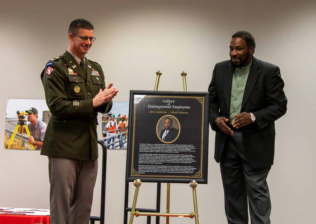 A white man in an Army colonel's uniform claps beside a Black man in a green shirt and black suit. Between them in a framed portrait of the Black man with gold detailing on a gold easel.