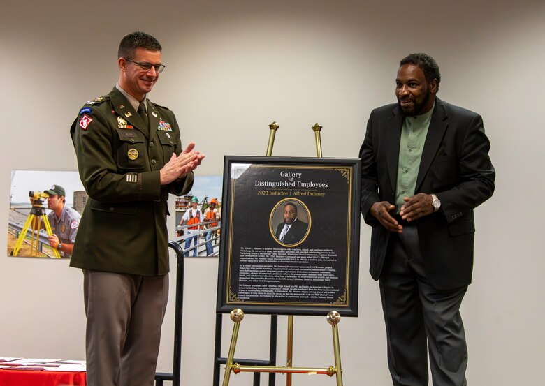 A white man in an Army colonel's uniform claps beside a Black man in a green shirt and black suit. Between them in a framed portrait of the Black man with gold detailing on a gold easel.