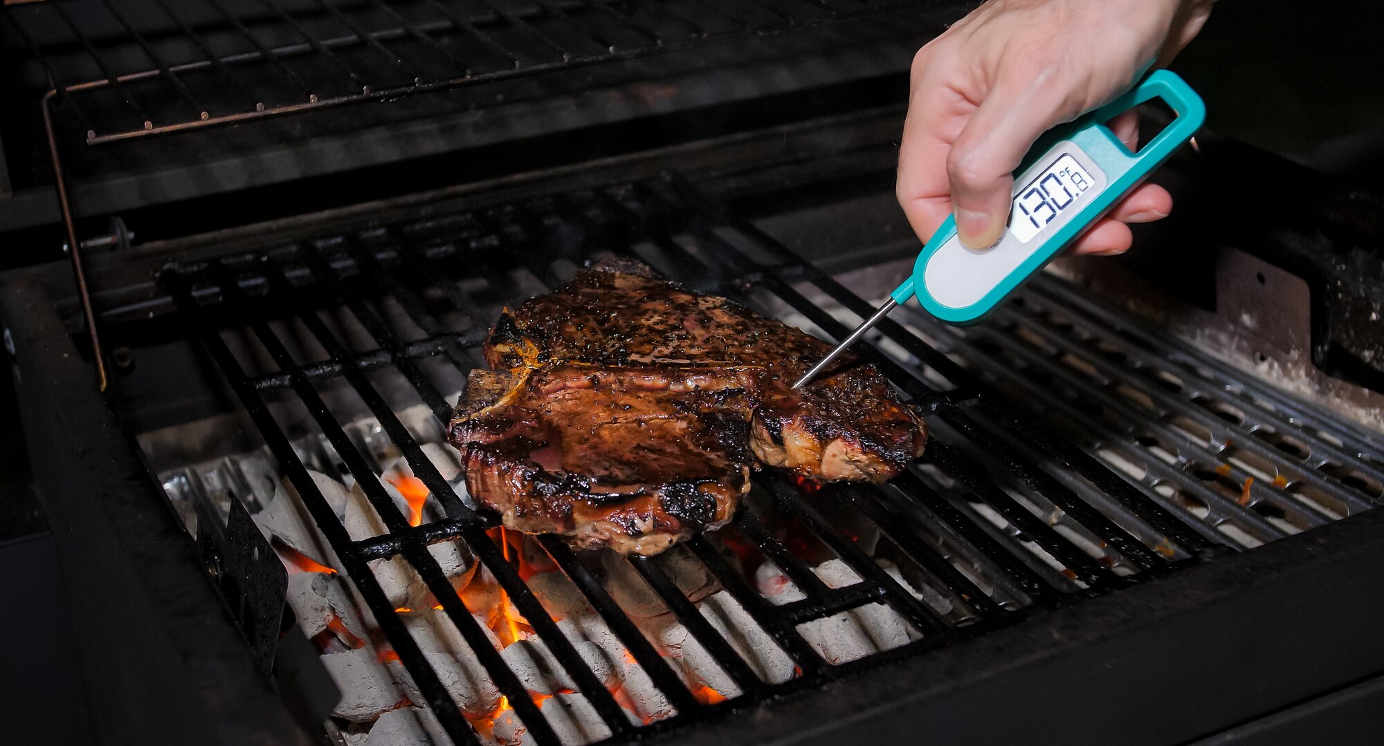 Photo of meat on a grill with a hand checking the temperature using a thermometer.