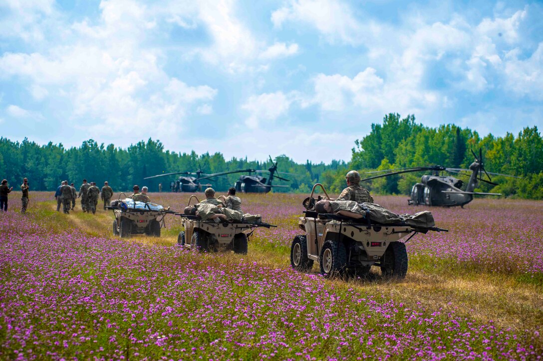 Guardsmen walk in front of other guardsmen transporting simulated patients on three military vehicles in a field of wildflowers next to three parked helicopters.