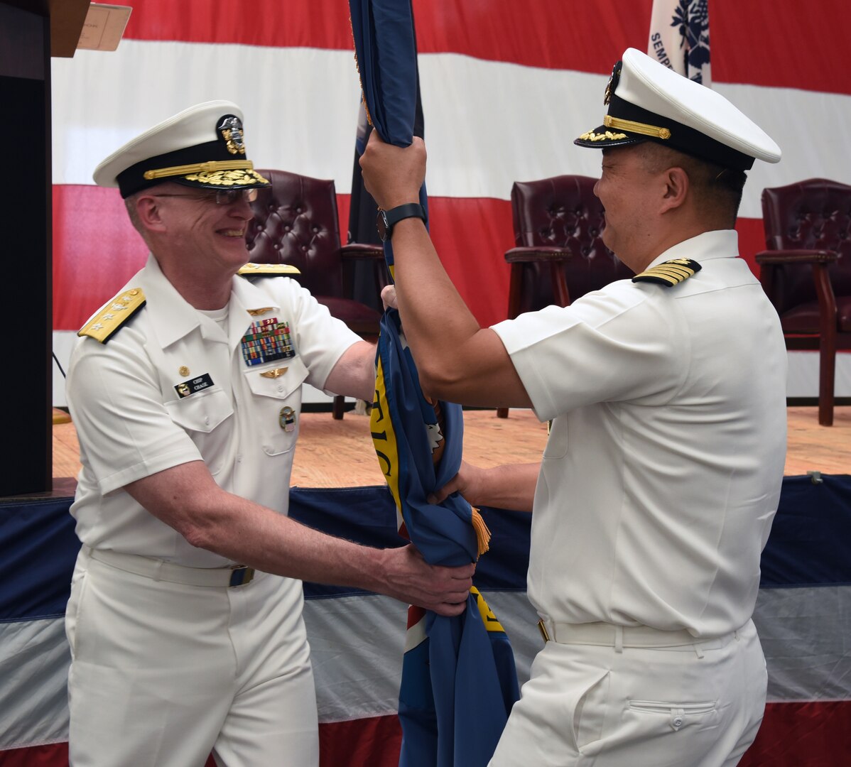 Photo is of two men in Navy white uniforms holding a large flag between them.