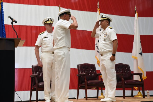 Three men in Navy white uniforms standing on a stage with a large American flag behind them. Two men are saluting.