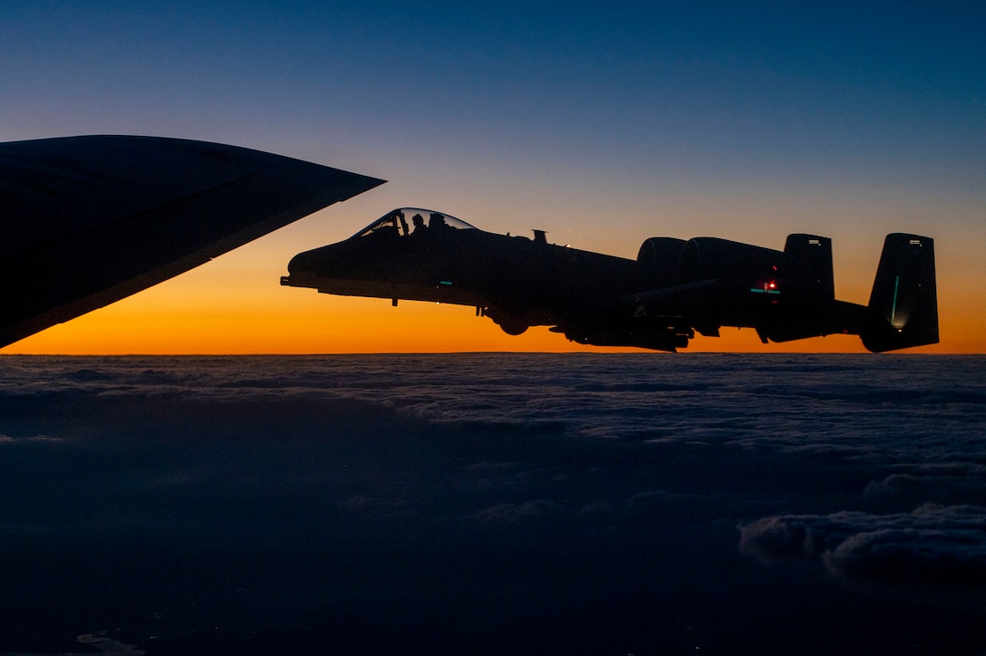 A military aircraft flies alongside the wing of another with a low-set sun in the background.