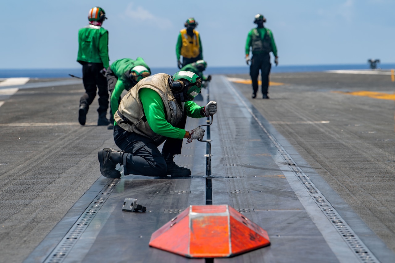 A sailor uses a tool to tighten bolts on the deck of a ship as others stand behind.