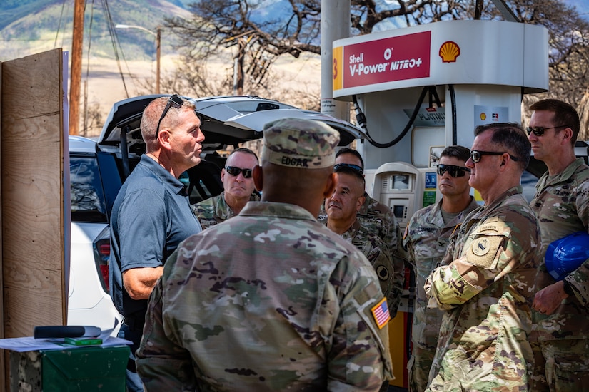 A person in civilian attire speaks to a group of service members in uniform gathered near a gasoline pump.