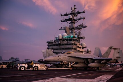 A Navy aircraft is moved on the flight deck of a ship with a cloudy, lavender sky in the background.
