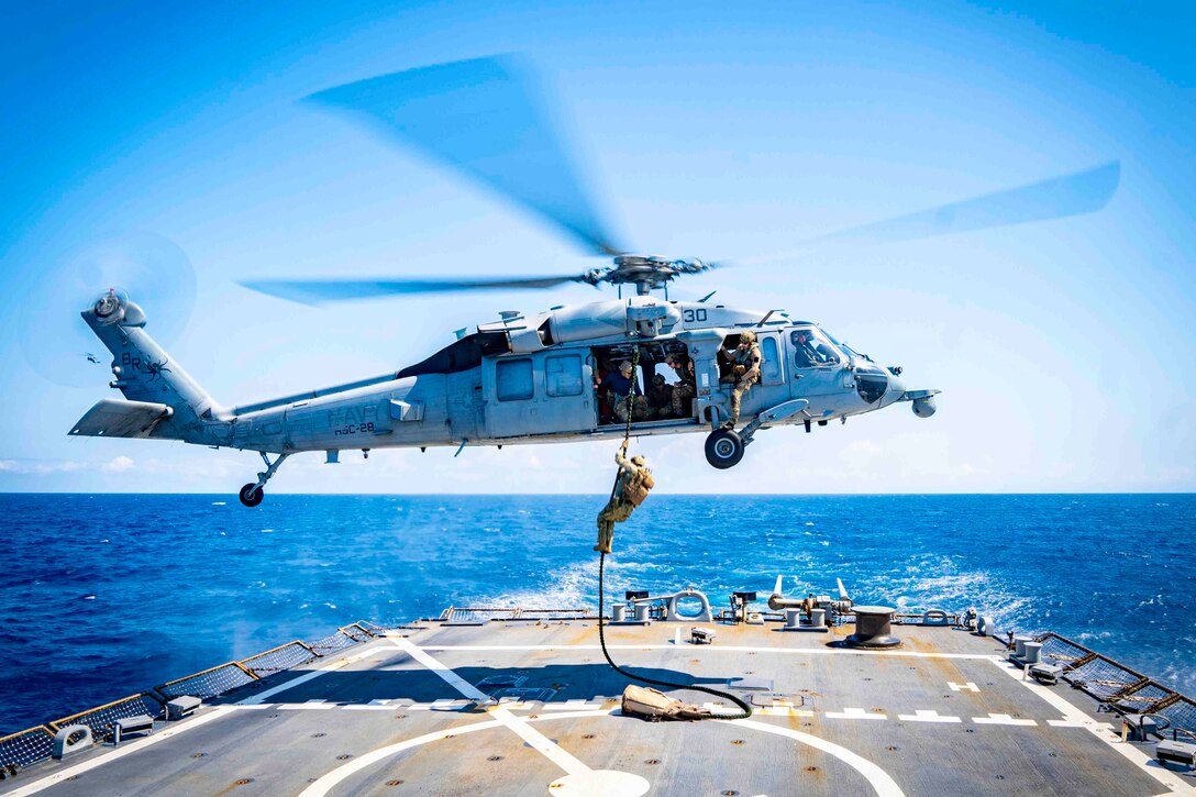 A sailor fast-ropes out of an airborne helicopter onto the deck of a ship.