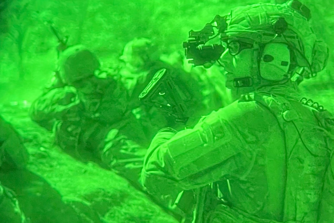 Soldiers are shown through a green night vision lens, lying on the ground while pointing their weapons as another stands in the foreground.