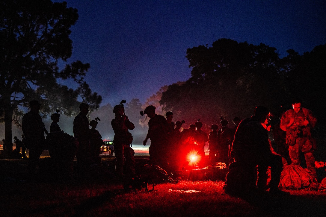 Soldiers congregate in a field at night while a red light illuminates the center of the photo.