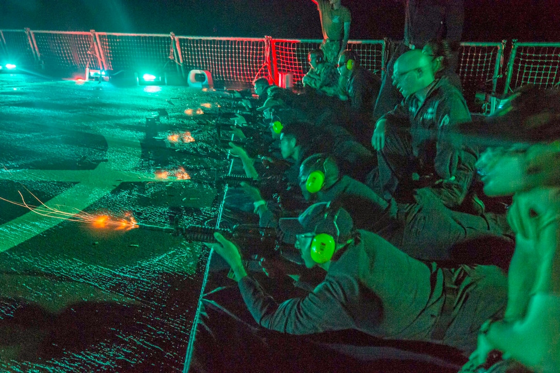 Sailors laying aboard a ship wearing protective gear fire weapons as fellow sailors kneel next to them and observe illuminated by colorful lights.