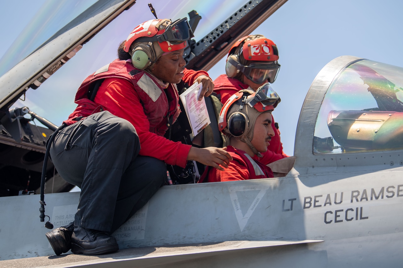 Three sailors sit in and around the cockpit of a jet.