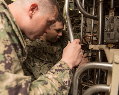Two men in camo uniforms look at the inner workings of a submarine