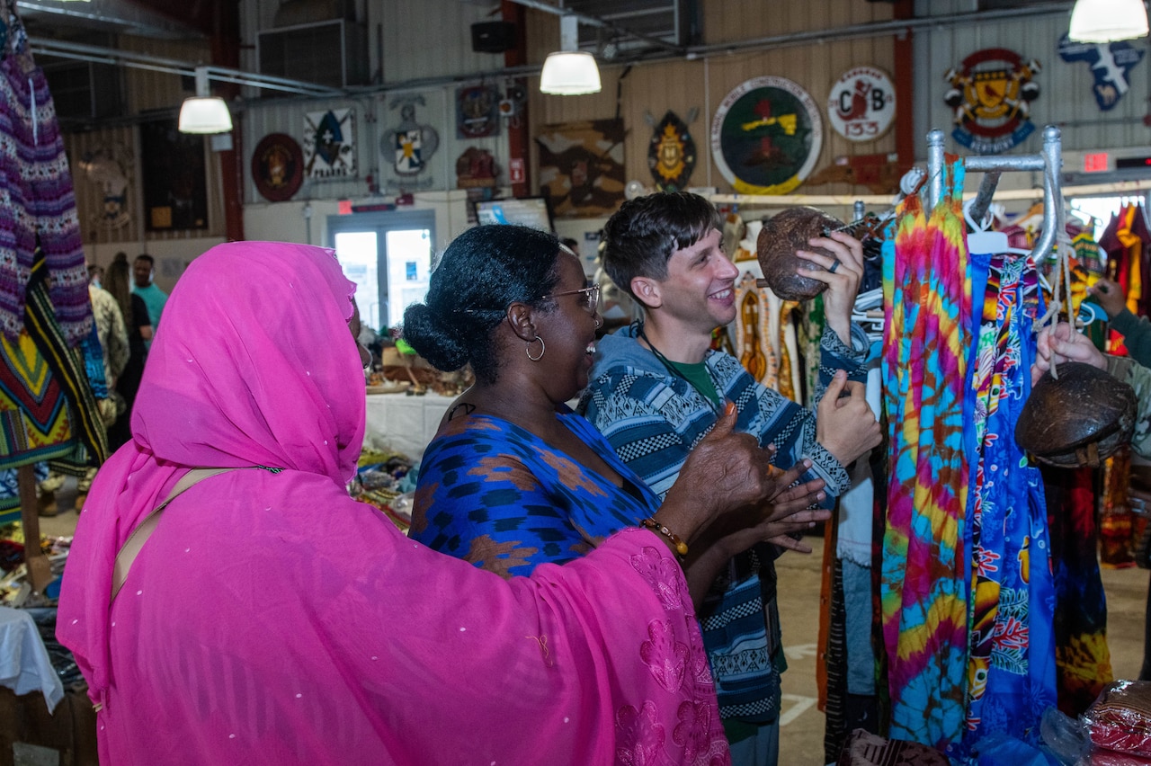 Two women observe as a man smiles while holding an art object at an indoor bazaar.