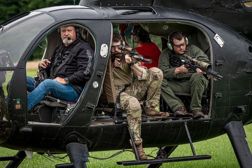 Two people aim weapons from a helicopter parked on the grass.