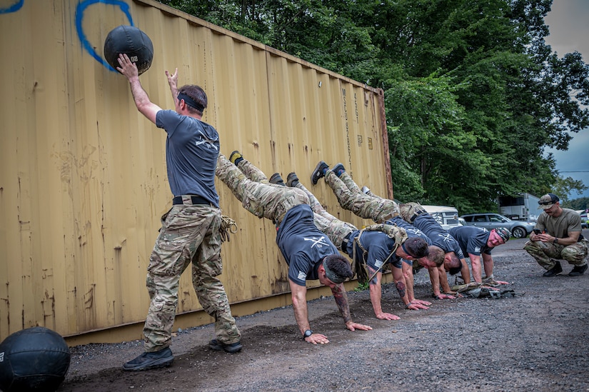 A soldier throws a medicine ball against a wall while team members hold an inverted position against it.